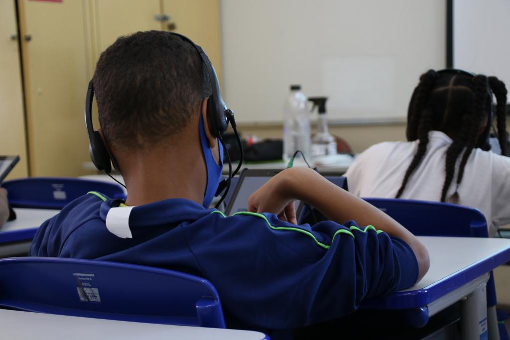  Foto por: Pedro Oliveira. #ParaTodosVerem. Descrição da imagem: Fotografia com uma criança de costas, usando uniforme, com fones de ouvido e fazendo uma atividade no tablet. 
