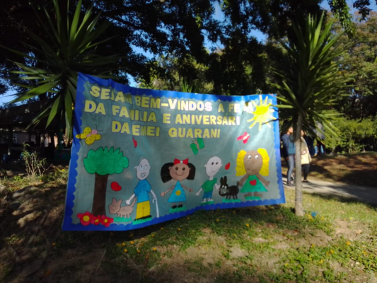 #paratodosverem Descrição: A fotografia contém um mural com os dizeres: “ Sejam bem-vindos à festa da família e aniversário da Emei Guarani”. Na parte de baixo, a retratação de uma família.