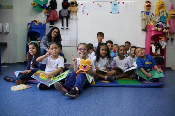 A imagem contém várias crianças uniformizadas, sentadas no chão da sala de aula de costas para o quadro e cada criança está segurando um livro.