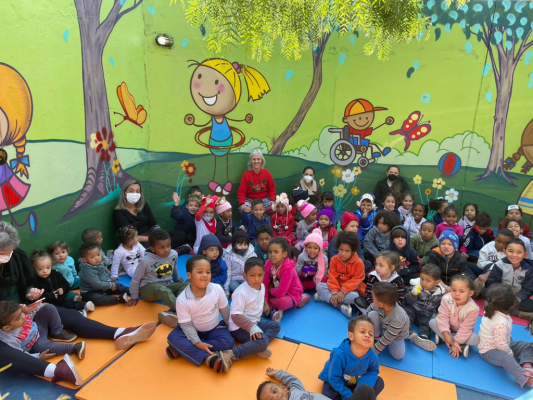 Muitas crianças sentadas no chão da escola, junto com 4 professoras e a autora do livro. Ao fundo, o muro pintado com árvores e crianças brincando. 