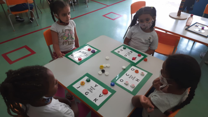 Fotografia de crianças vestindo uniformes brancos da PBH e desenvolvendo atividade com tampas de garrafas e jogo de palavras impresso. 