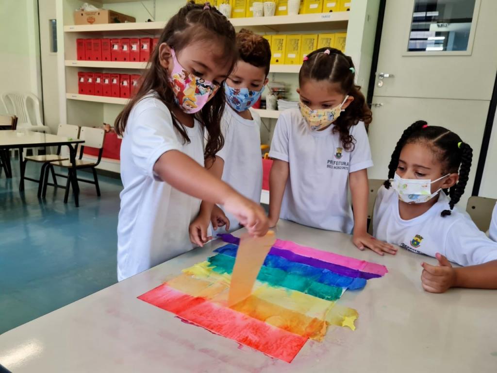  Fotografia de crianças usando uniforme da Rede Municipal de Belo Horizonte e trabalhando o grafismo com papéis coloridos nas cores: rosa, azul, roxo, amarelo, verde laranja e vermelho.