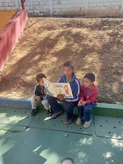#pratodosverem: fotografia colorida de alunos da EMEI União Comunitária lendo o mesmo livro juntos. Eles vestem blusas de frio, um usa chinelo e os outros dois calçam tênis. 