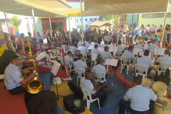 Fotografia de músicos da Guarda Municipal sentados com instrumentos musicais.Ao fundo, familiares e estudantes em pé assistindo à apresentação.