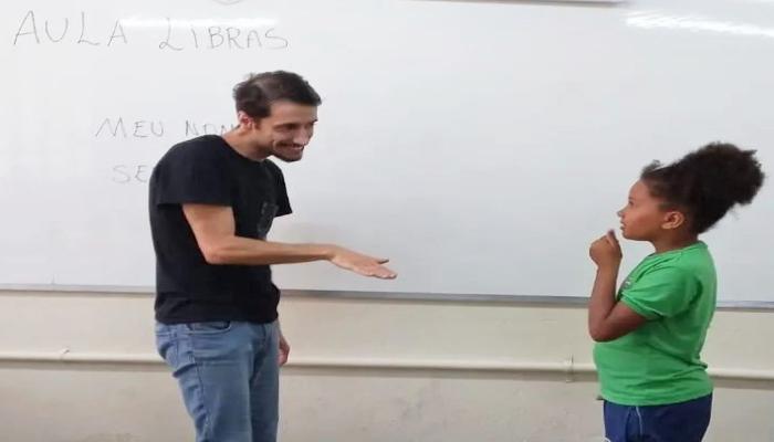 #paratodosverem Descrição: fotografia colorida de um professor e uma aluna conversando em Libras, de pé, na frente do quadro branco. Foto por: M. Rui da Costa Val.