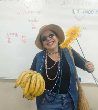 #paratodosverem descrição: fotografia colorida de Maricota, personagem rural. Ela usa roupa azul e preta, usa óculos escuros e colares, está em pé, carrega uma bolsa e segura um cacho de bananas e dois girassóis nas mãos.