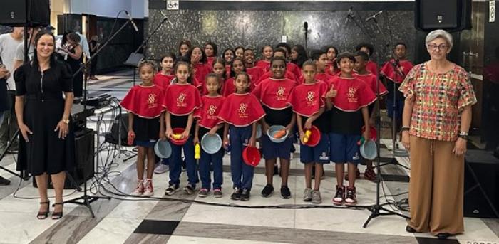 #paratodosverem descrição: fotografia colorida de cerca de 20 estudantes da EM Maria Silveira. Eles estão de pé, disposos em três fileiras, vestem blusa azul, calça do uniforme escolar e pelerine vermelho. Do lado direito do grupo, sorridente, está a professora Cecília, e do lado esquerdo, a professora Dóris Matia Fonseca Ferreira, coordenadora do Programa Escola Integrada da instituição. Foto: Beatriz Rodrigues