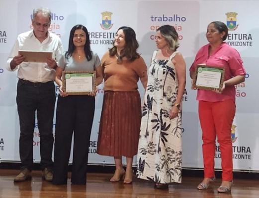 Legenda: Belo Horizonte recebe prêmio pelos 30 anos da Política Alimentar e Nutricional da cidade. #paratodosverem descrição: fotografia colorida de cinco pessoas em cima de um palco, recebendo o prêmio. Elas estão sorrindo e três delas seguram uma placa. Da esquerda para a direita estão: Afonso Barbosa (secretário adjunto de educação), Renise Britto (vice-diretora da EM João Pinheiro), Darklane Dias (subsecretária de seguraça alimentar e nutricional), Adilana Rocha (diretora de assistência alimentar) e Patrícia Cruz (cantineira da EM Aurélio Pires). Foto: Beatriz Rodrigues