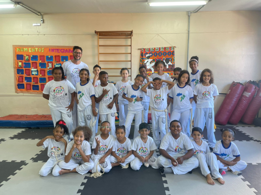 #paratodosverem: fotografia colorida em plano aberto da equipe de capoeira da EM Wladimir de Paula Gomes, uniformizada, posando em uma sala com tatames e equipamentos de luta.