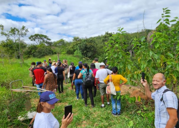 #paratodosverem: fotografia colorida que mostra um campo verde. O céu está nublado, mas é possível ver o azul claro, sinalizando que a foto foi tirada durante o dia. Dezessete pessoas adultas estão de costas, como se estivessem andando para a frente. Uma mulher e um homem estão em primeiro plano. A mulher utiliza um boné roxo e blusa branca, enquanto o homem veste camisa xadrez azul e branca e porta uma mochila. O rosto de ambos aparece parcialmente na foto e seguram celulares, registrando o momento.