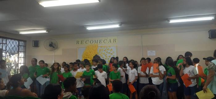 #paratodosverem: fotografia colorida tirada em plano aberto de uma sala de aula. Os(As) estudantes estão em frente às mães e aos pais, com papéis coloridos na mão, para recitarem a música citada no texto.