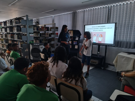 #paratodosverem: fotografia colorida de um estudante participando da apresentação contra o bullying. Em uma sala escura, diversos(as) estudantes assistem a uma apresentação e um deles está na frente de todos. Atrás dele, uma televisão projeta uma imagem com os dizeres “Através do amor-próprio (sentimento de dignidade, respeito que cada um tem por si mesmo).”