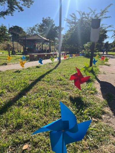 #paratodosverem: fotografia colorida do parque enfeitado com cataventos coloridos. A foto, tirada muito próxima ao chão, centraliza uma faixa de grama decorada com cataventos azuis, verdes, vermelhos e amarelos nas suas duas extremidades. Ao fundo, é possível ver os brinquedos e a estrutura do parque destinada a festa