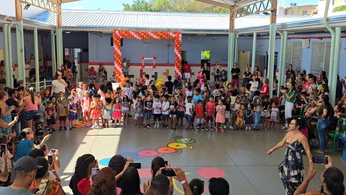 #paratodosverem: fotografia colorida do pátio da escola repleto de pessoas comemorando a Festa da Família. Ao fundo, dezenas de crianças estão reunidas em filas enquanto diversos adultos ao seu redor observam e fotografam o momento com o celular.