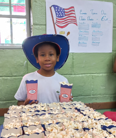 #paratodosverem: fotografia colorida de um menino participando da exposição da pipoca como alimento típico dos Estados Unidos. Atrás de uma mesa contendo vários pacotes de pipoca, a criança segura dois deles enquanto usa um chapéu azul e vermelho. Atrás dele, pregada na parede, está uma cartolina branca com a bandeira do país e um texto escrito com o título “Comidas Típicas dos Estados Unidos - 6º ano”.