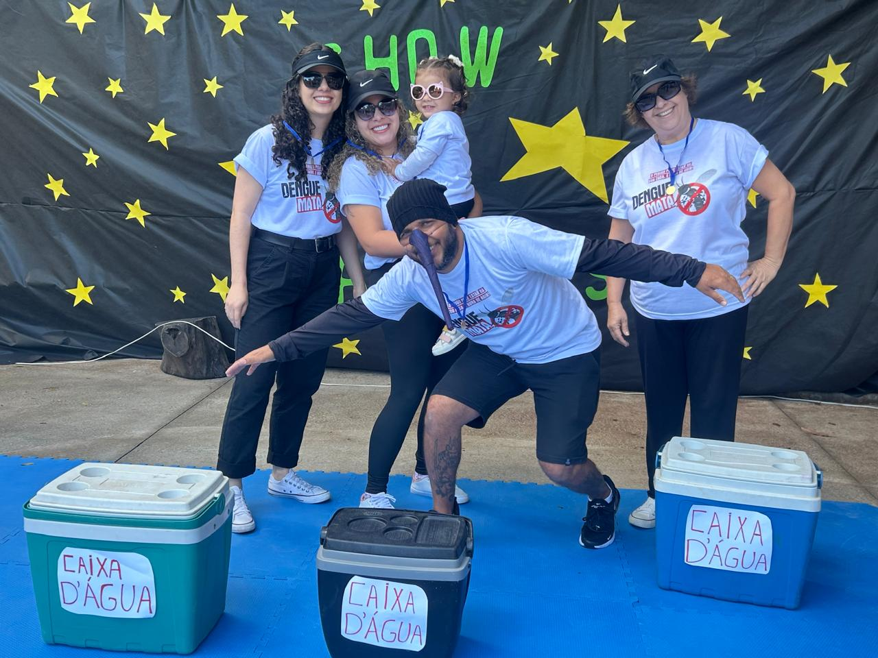 #paratodosverem: um grupo de cinco pessoas posa sorridente em frente a um painel preto decorado com estrelas amarelas e a palavra "SHOW" em verde. Todos vestem camisetas brancas com a frase “Dengue Mata” e o símbolo de combate ao mosquito da dengue. Três caixas térmicas estão no chão. Uma das mulheres segura uma criança de óculos escuros no colo, enquanto um homem faz pose divertida.