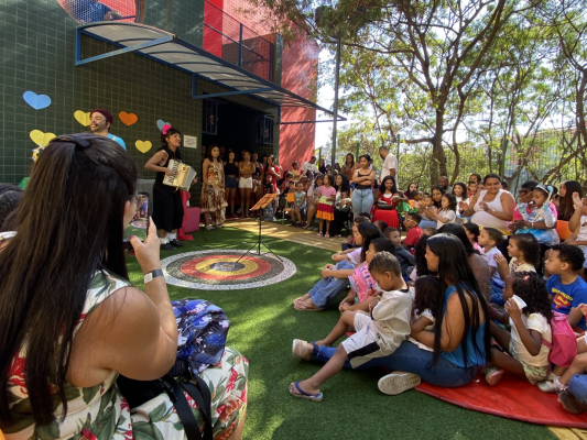 #paratodosverem: em um pátio escolar, um grupo grande de crianças e famílias assiste a uma apresentação musical. No centro, dois artistas tocam sanfona e cantam. As crianças estão sentadas no chão, atentas, enquanto os adultos assistem em pé ou sentados nas laterais. Uma mulher em primeiro plano registra o momento com o celular. 