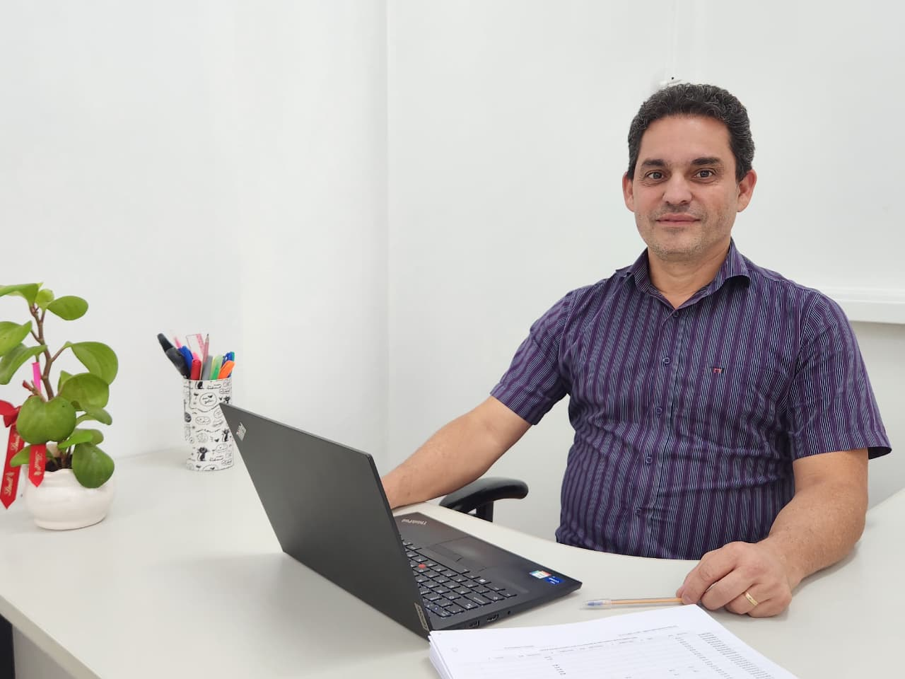 Homem branco, sentado à mesa, usando camisa social listrada com tons em azul e roxo, utilizando notebook. 