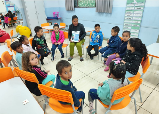 #paratodosverem: Crianças estão sentadas em círculo em uma sala de aula, ocupando cadeiras laranjas. Ao centro, uma educadora segura um jornal ou revista aberta, mostrando para o grupo. Todas as crianças estão atentas à atividade. Ao fundo, há mesas, cadeiras e materiais escolares.