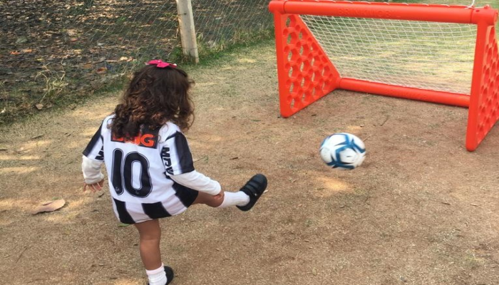 #paratodosverem #Descrição: fotografia colorida de uma garota usando camisa típica de clubes de futebol na cor preta e branca e chutando uma bola branca em direção ao gol de traves alaranjadas. Foto: acervo Emei Sarandi.