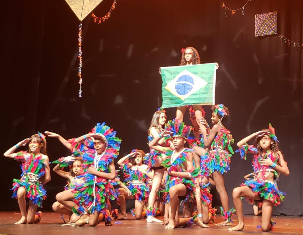 Dez crianças com fantasias de índio fazem apresentação em um palco e uma delas segura uma bandeira do Brasil