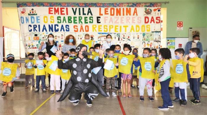 #paratodosverem Descrição: Fotografia de crianças da Emei Vila Leonina usando coletes amarelos para a campanha de prevenção à dengue e um homem entre elas, vestido de Mosquito Aedes aegypt Foto: Acervo Emei Vila Leonina