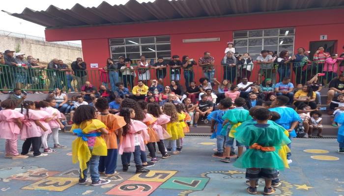 #paratodosverem Descrição: fotografia colorida com diversas pessoas observando a apresentação de crianças, que estão em fila usando vestes em tons coloridos. Foto: Escola Municipal Monteiro Lobato. 