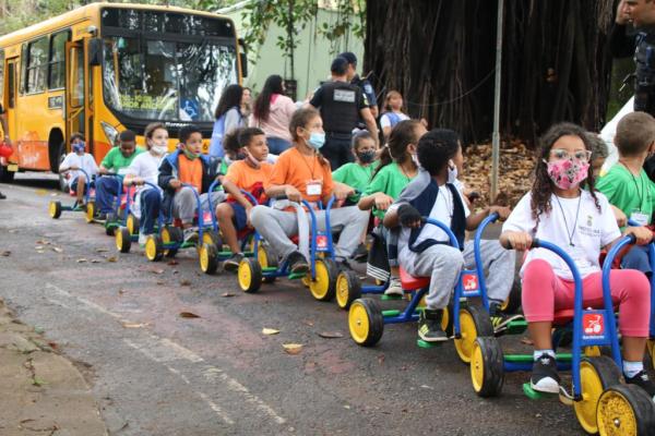 #paratodosverem Descrição: Fotografia colorida de crianças uniformizadas com as roupas da Rede municipal de Educação de BH, sentadas em triciclos infantis. Ao fundo, ônibus municipal na cor amarela e membros da Guarda Municipal.