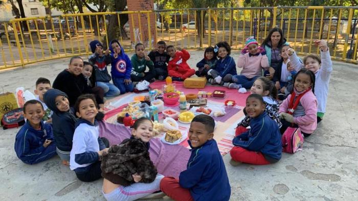 #paratodosverem Descrição: Fotografia colorida de estudantes sentados no chão e alimentos saudáveis sobre um tapete colorido em tons de rosa e roxo. Foto por: Professora Nara Ferreira.