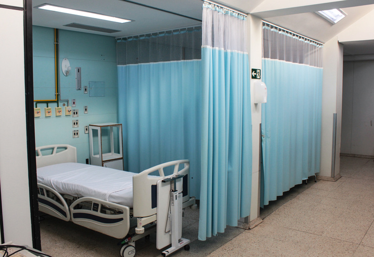 Hospital Odilon Behrens otimiza acesso a leitos e reduz tempo de espera com novo modelo de gestão Leito de hospital com uma cama hospitalar branca vazia e aparelhos de apoio, um leito com cortinas azuis abertas e outro com cortinas fechadas.