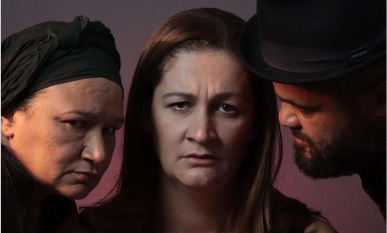 Espetáculo no Teatro Marília reflete sobre machismo e violência contra a mulher Um close-up dramático de três atores em um estúdio com iluminação lateral quente. No centro, uma mulher de cabelos castanhos longos olha severamente para a câmera. À esquerda dela, uma mulher com um lenço de cabeça preto de textura grossa olha para a mulher central com desconfiança. À direita, um homem de barba cheia e chapéu-coco preto a observa fixamente de perfil. As três figuras estão muito próximas umas das outras contra um fundo escuro e desfocado, evocando tensão psicológica