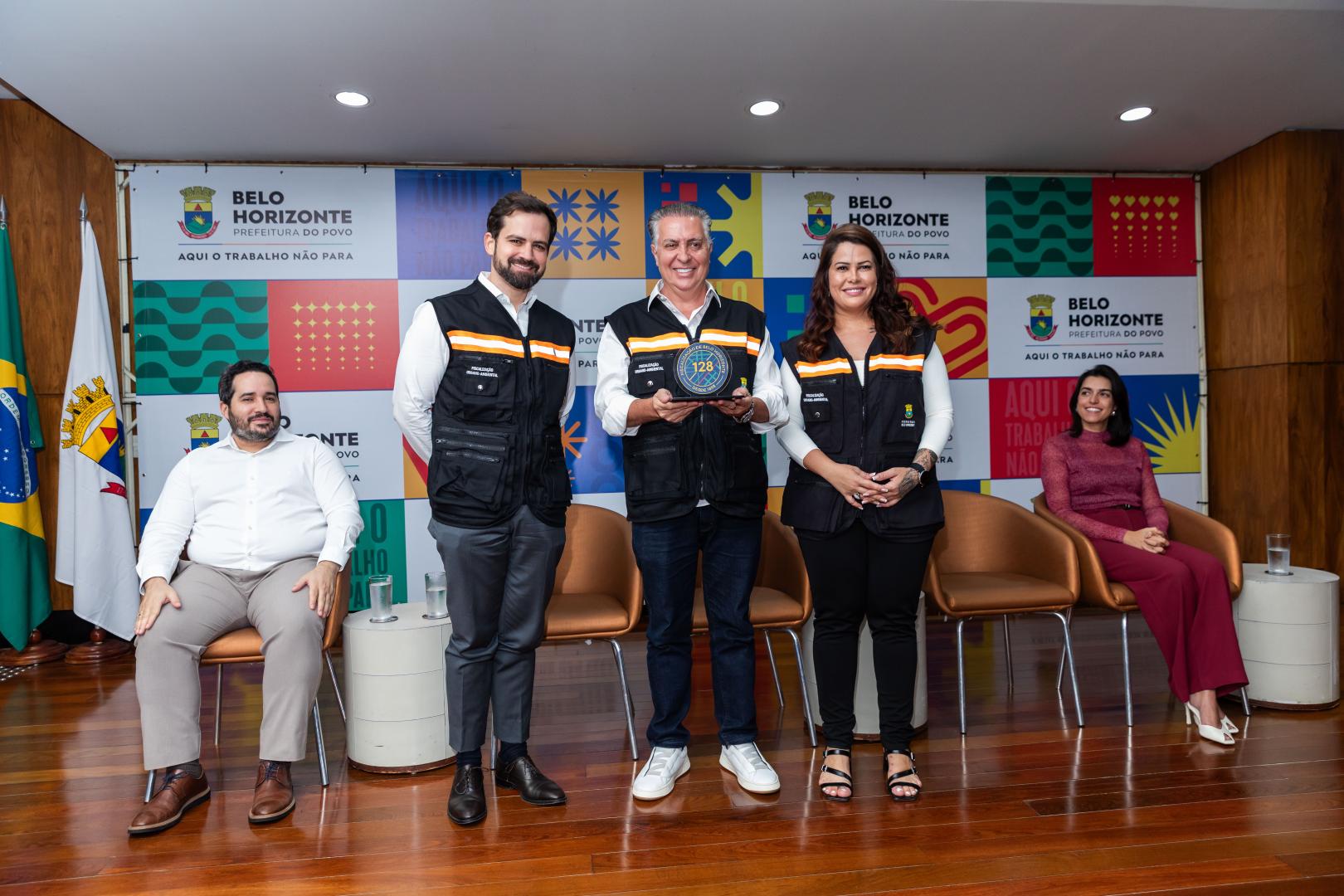 BH homenageia parceiros nos 128 anos da carreira de Fiscalização A foto registra um momento de reconhecimento institucional em um auditório da Prefeitura de Belo Horizonte. Ao centro, o prefeito Álvaro Damião segura uma placa comemorativa com o número 128, acompanhado de outros três participantes que estão em pé, vestindo coletes pretos com detalhes em laranja e camisas brancas. Duas pessoas estão sentadas nas extremidades, uma de cada lado do grupo. Ao fundo, há um painel colorido com elementos gráficos e a identificação da Prefeitura de BH, além de bandeiras laterais.