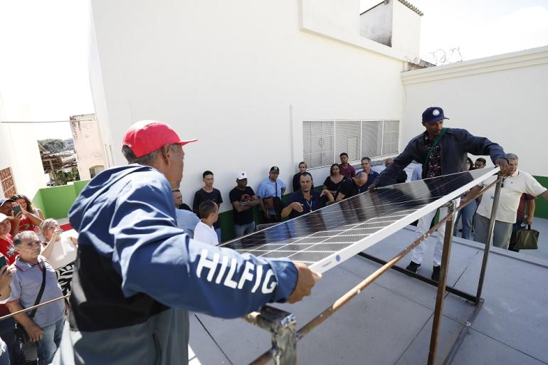 Dois homens no centro da foto segurando uma placa fotovoltaica, pessoas ao sentadas e em pé ao redor deles, atentos ao o que está acontecendo. 