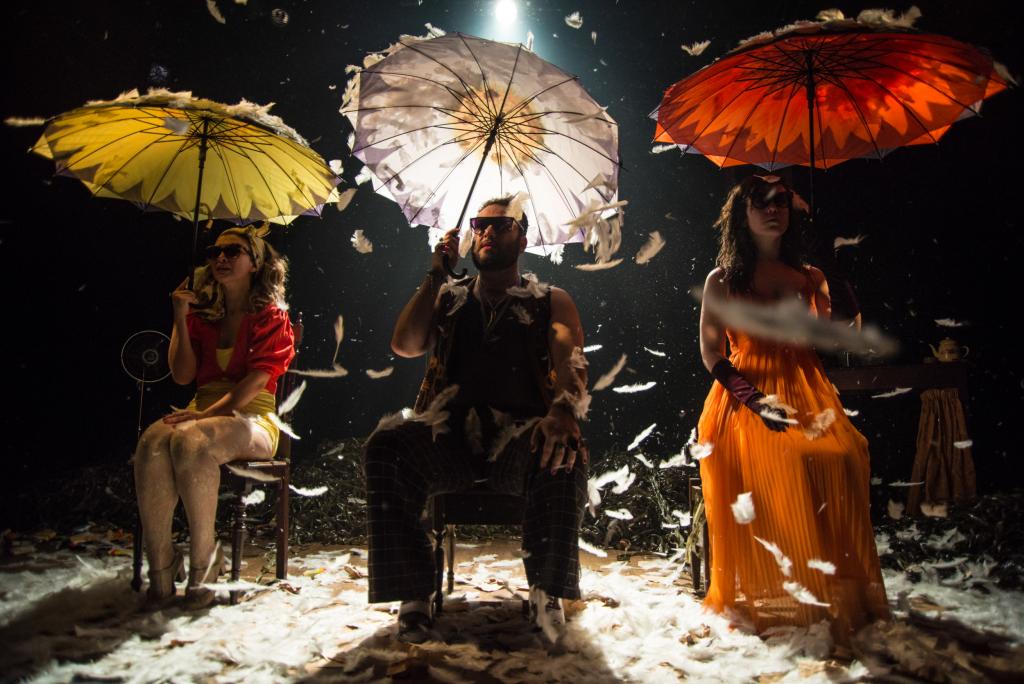 Três pessoas, sentadas em três cadeiras, seguram guarda chuvas em cima de um palco, com iluminação direta neles.