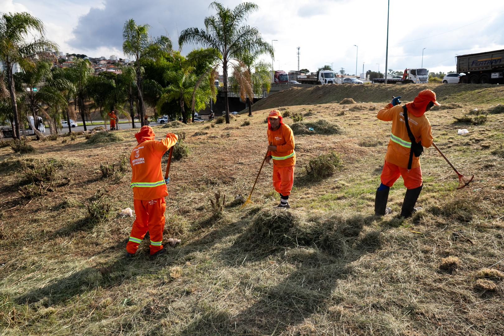 PBH amplia manutenção e conservação do Novo Anel Pessoas uniformizadas e com equipamentos de proteção realiza roçagem da vegetação às margens de uma via, utilizando uma roçadeira. Ao fundo, há ônibus, veículos, placas de trânsito e uma área urbana com moradias.