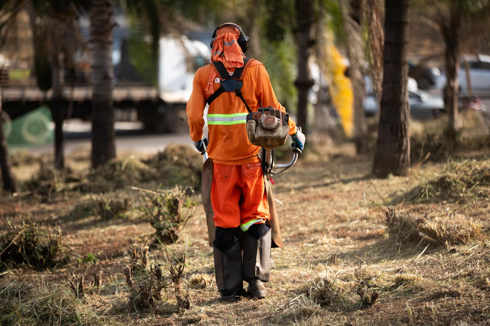PBH amplia manutenção e conservação do Novo Anel Pessoa uniformizada e com equipamentos de proteção realiza roçagem da vegetação às margens de uma via, utilizando uma roçadeira. Ao fundo, há ônibus, veículos, placas de trânsito e uma área urbana com moradias.