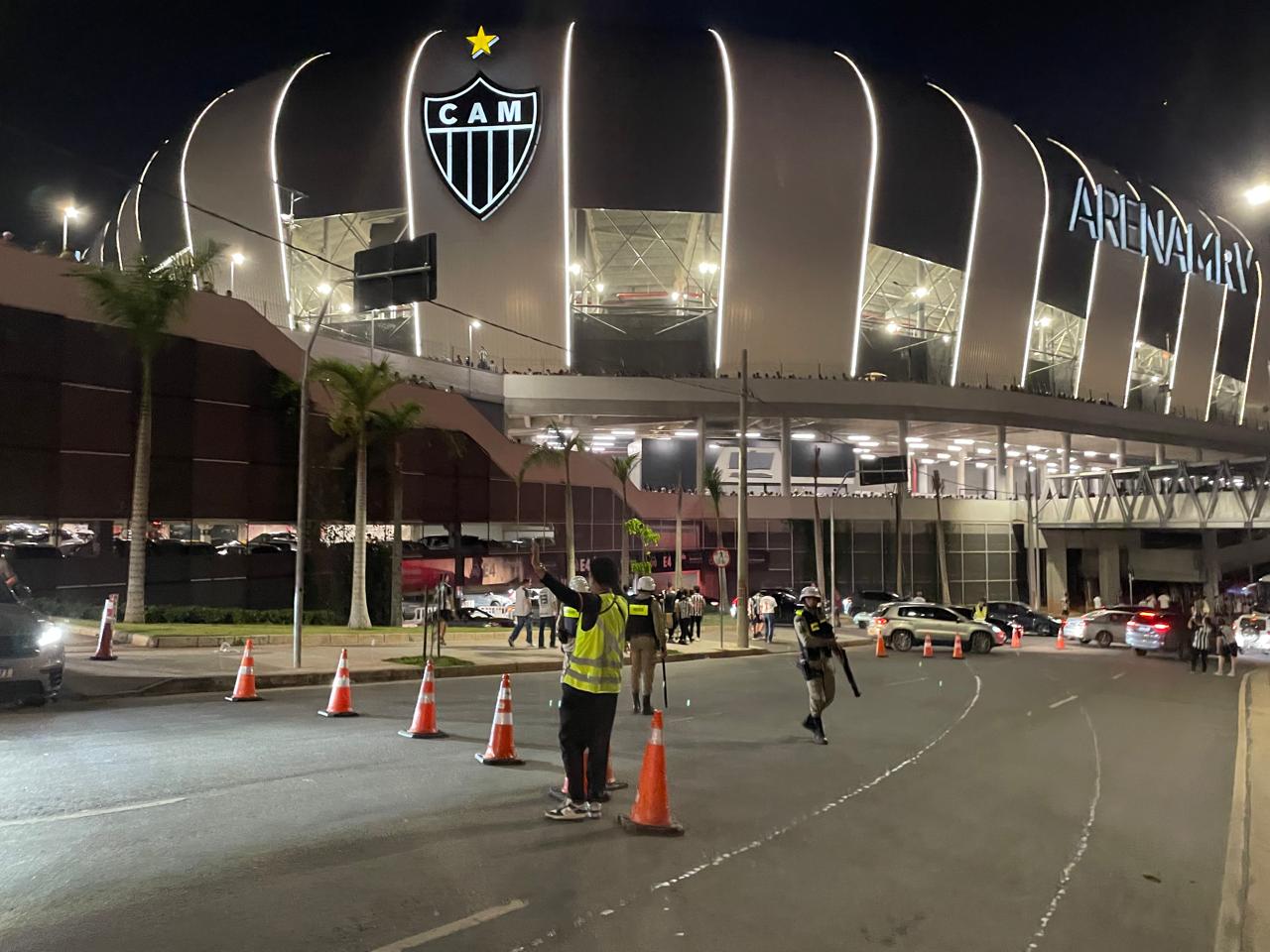 Operação de trânsito para Atlético x Coritiba na Arena MRV, neste sábado (17) Foto noturna da Arena MRV, estádio do Atlético Mineiro, com a fachada iluminada e o escudo do clube em destaque. Na rua em frente, agentes de trânsito organizam a circulação de veículos com o auxílio de cones.