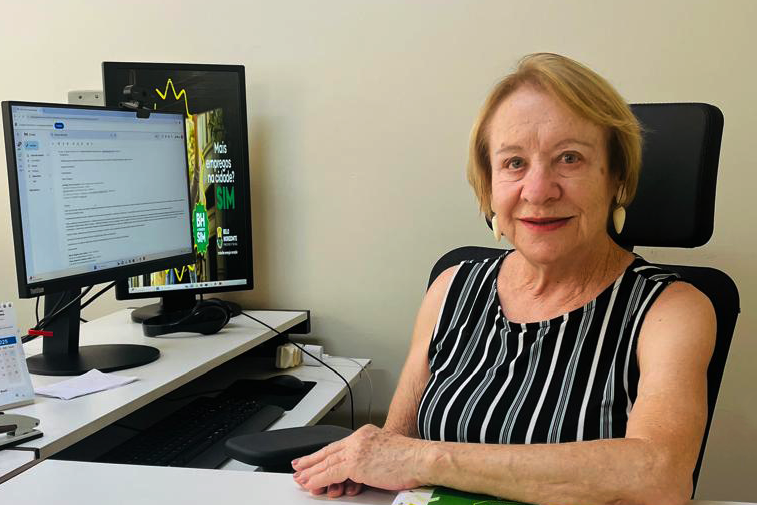 mulher de meia idade, cabelos curtos e loiros vestindo camiseta preta listrada e sentada em frente a um computador