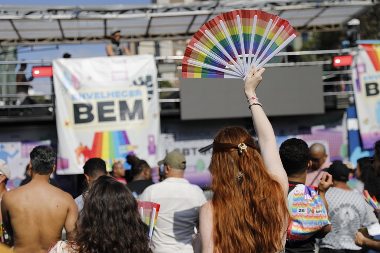 Prefeitura apoia a realização da 27ª Parada do Orgulho LGBTQIAPN+ de Belo Horizonte Pessoas de costas para o palco participam da Parada LGBT. Em destaque, uma mulher branca, ruiva, segura um leque nas mãos