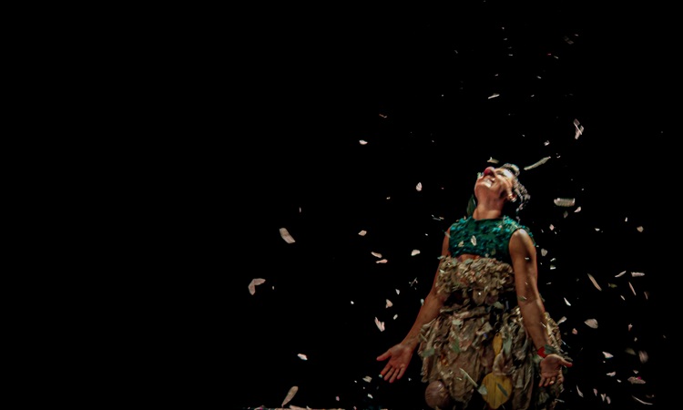 Teatro Raul Belém Machado recebe espetáculo circense sobre ancestralidade e cura Uma artista veste um traje complexo com tema natural, composto por um top verde-esmeralda e uma saia de camadas de folhas secas e materiais naturais. A artista está com a cabeça inclinada para trás, olhando para cima com uma expressão alegre, e com os braços erguidos e estendidos para os lados. Papéis prateados e confetes iridescentes caem em torno deles contra um fundo completamente preto. A iluminação dramática destaca a textura do traje e o movimento dos confetes.