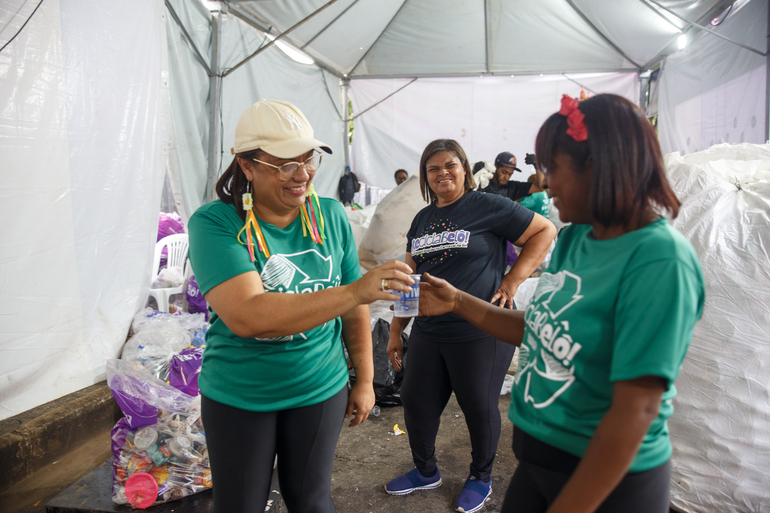 Arraial de Belô terá coleta seletiva e esquema especial de limpeza no Mineirinho Três mulheres, duas com camisa verde de frente uma para a outra e outra com uma camisa preta do ReciclaBelô, todas dentro de uma barraca branca cercadas por materiais para reciclagem.