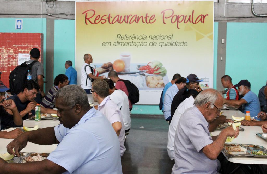 Cerca de quinze cidadãos sentados em duas mesas, almoçando, em uma sala com um banner do Restaurante Popular ao fundo. 