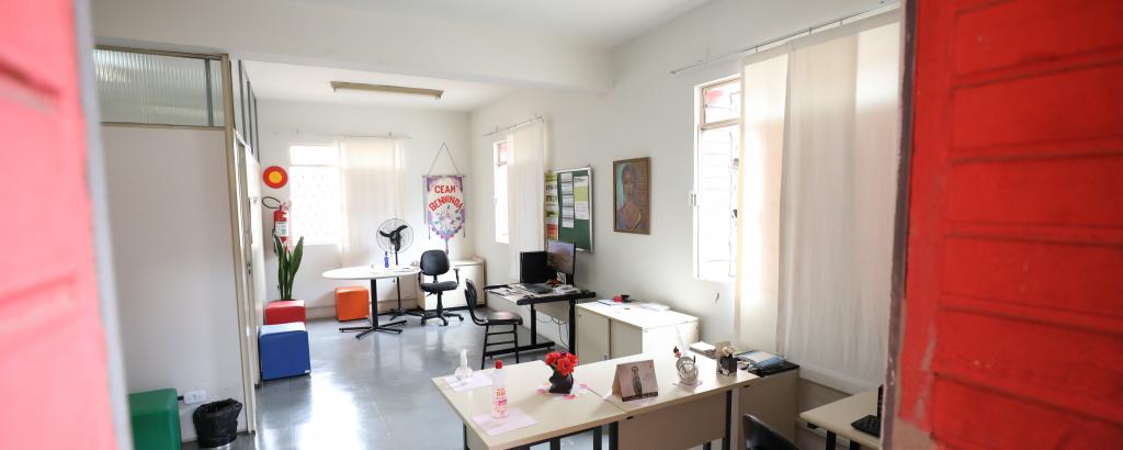 Foto de uma sala do Centro de Atendimento à Mulher Benvida, com mesas e cadeiras para atendimento. Em uma parede ao fundo há um standart com o escrito BENVINDA