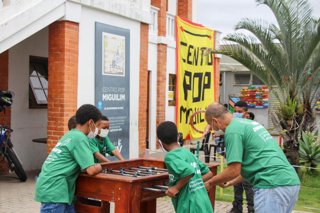 Crianças usando camisas verdes jogam futebol de mesa em frente ao prédio do Centro Pop Miguilim