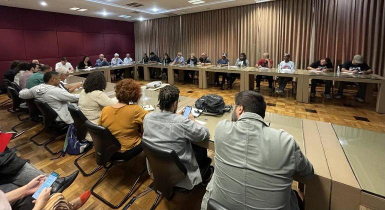 Foto de uma sala de reuniões com cerca de 50 pessoas sentadas ao redor de grande mesa com formato de U