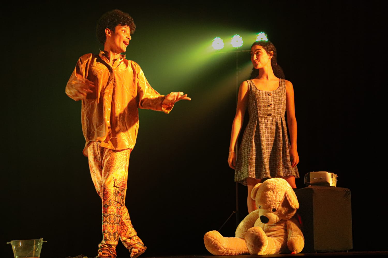 Teatro Marília recebe duas apresentações da comédia “Somos Tão Jovens” Dois atores num palco iluminado por luzes esverdeadas: à esquerda, um rapaz com camisa folgada e calças estampadas gesticula alegremente; à direita, uma jovem de vestido xadrez observa-o, perto de um urso de pelúcia gigante sentado no chão.
