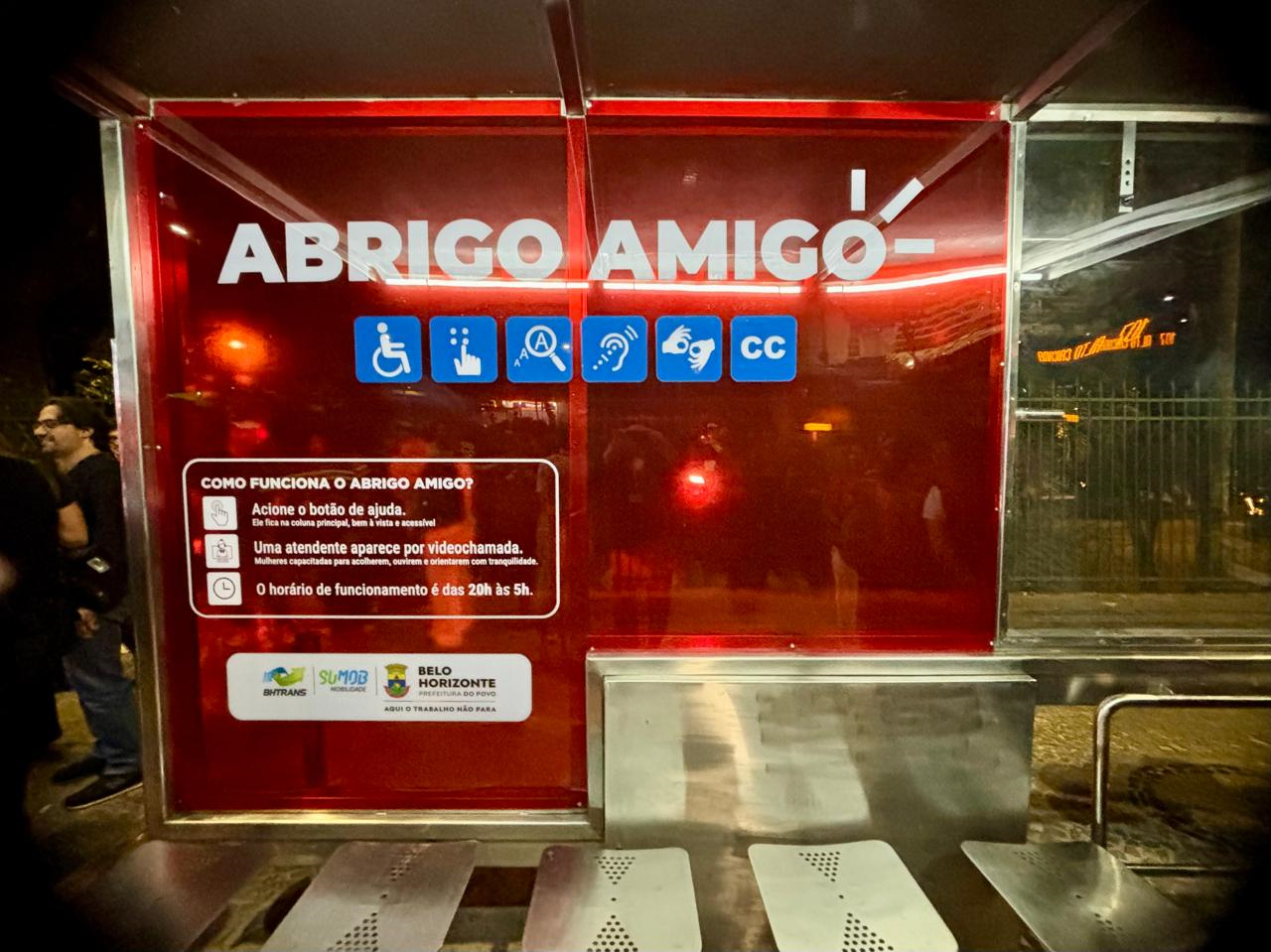 Painel luminoso vermelho em um ponto de ônibus noturno com o título "ABRIGO AMIGO" e ícones de acessibilidade na cor azul, logo embaixo do título.