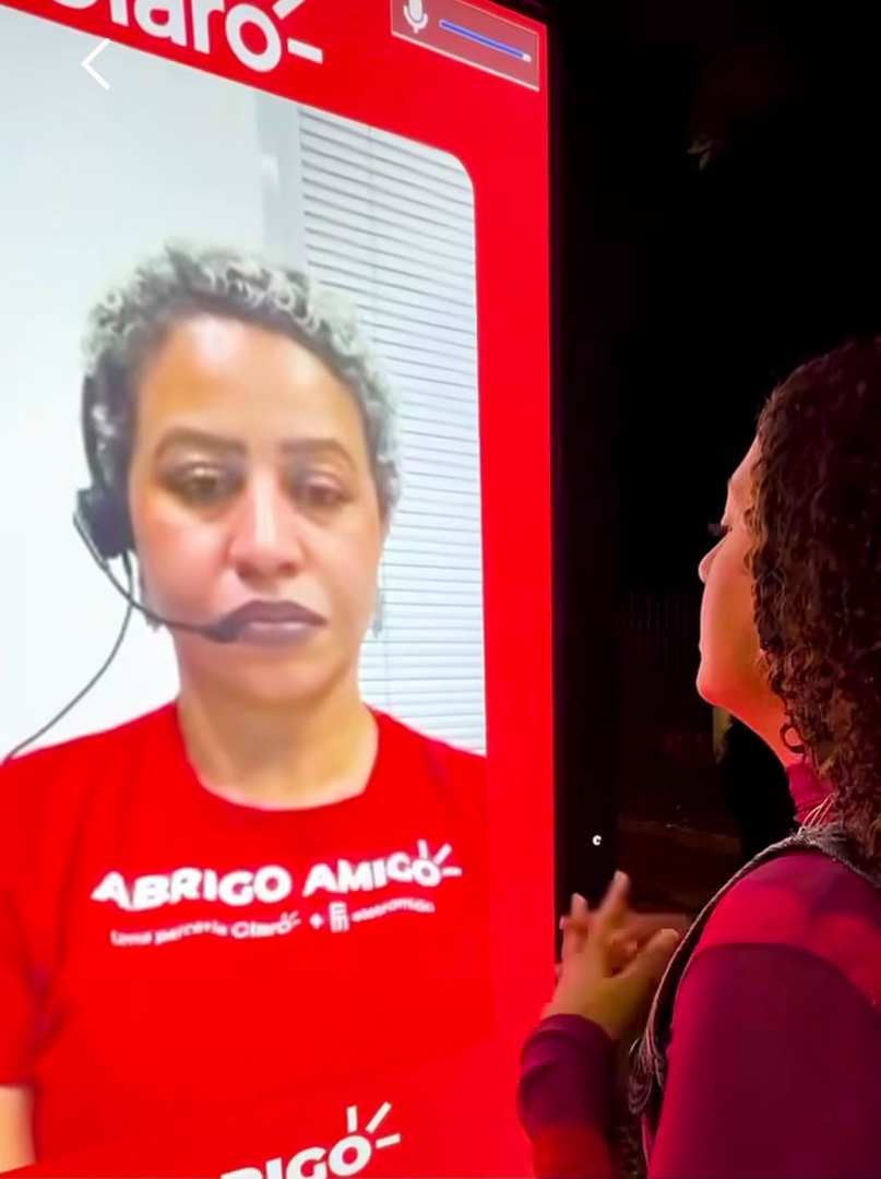 Pessoa observa um painel digital do Abrigo Amigo, que exibe na tela uma mulher usando headset durante uma videochamada do atendimento do Abrigo Amigo.
