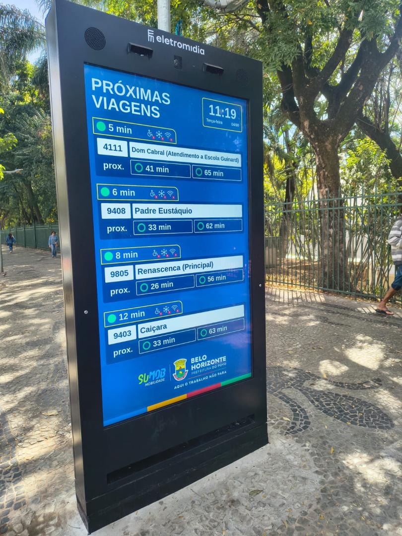 Painel digital do serviço Próxima Parada, instalado em uma calçada arborizada, exibindo em tempo real as próximas linhas de ônibus, destinos e previsão de chegada.