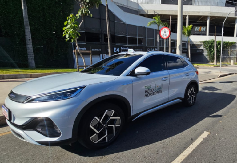 Após mais de 20 anos, serviço de táxi é integrado entre BH, Confins e Lagoa Santa Um carro branco com a identificação “Táxi Belo Horizonte” está estacionado em uma rua, em frente a um prédio. Ao fundo, há árvores, placas de trânsito e a entrada do estacionamento.